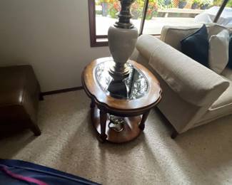 Traditional wooden oval end table with a mirrored glass insert top, accompanied by a ceramic and brass table lamp with a pleated shade.