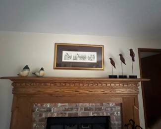 Framed waterfowl art print in a gold tone frame hanging above a wooden fireplace mantle displaying carved bird figurines.