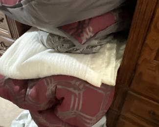 Stack of contemporary bed linens including a dusty rose patterned comforter, decorative throw pillows, sheets, and a white blanket.