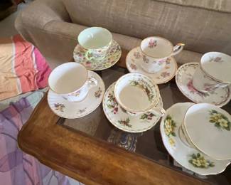 Collection of six fine bone china teacups and matching saucers with various floral patterns, displayed on a glass-top wooden coffee table.