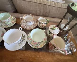 Collection of vintage floral bone china teacups, matching saucers, creamer pitchers, and a covered sugar bowl displayed on a wooden coffee table alongside a decorative hourglass.