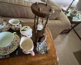 Collection of tabletop decor including an hourglass with brass and wood accents, floral china teacups and creamers, and a clear glass leaf dish.