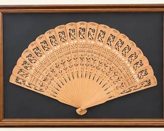 Decorative carved folding fan displayed in a rectangular wood-tone frame against a dark background. Intricate pierced floral-style detailing.