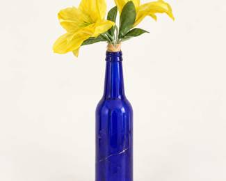 Cobalt Blue Glass Bottle Vase ◆ Deep blue glass bottle holding artificial yellow lilies and green foliage. Decorative floral arrangement.