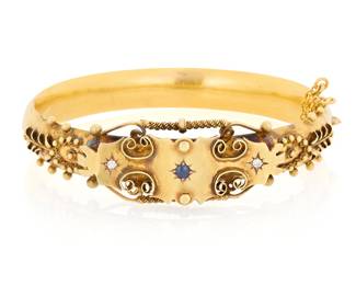 1044 A Victorian Gold, Sapphire and Diamond Bangle Of a hinged design featuring scrolling gold motifs and granulation accents, centering two round diamonds and a round sapphire  Inner circumference: 6 3/4 in. 15.8 grams gross Estimate: $1,000 - $1,500