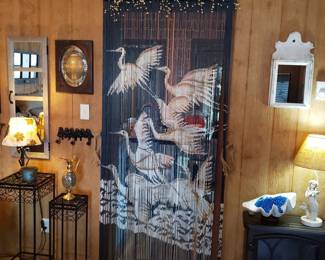 Vintage doorway curtain featuring a painted motif of flying egrets, flanked by various home decor items including metal plant stands, small lamps, and wall mirrors.
