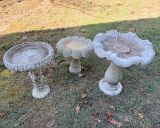 Three Birdbaths – Tree Trunk and Flower Designs Three concrete-look birdbaths: a tree trunk-style pedestal bath, a scalloped flower basin and a large petal-edge flower birdbath. Garden birdbaths for a yard or patio. Weathering consistent with outdoor use.