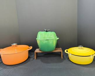 Enameled Dutch Ovens – Orange Oval, Le Creuset Yellow Oval and Green Bell Pepper Three enameled cookware pieces: an orange oval Dutch oven, a yellow oval Dutch oven with an embossed Le Creuset lid, and a green bell pepper-shaped covered pot. Colorful enameled cookware for collectors and home cooks.