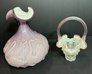 Lavender Swan Embossed Glass Pitcher and Iridescent Ruffled Basket A lavender glass pitcher with embossed swans and cattails and a white ruffled rim, paired with an iridescent glass basket with a ruffled edge and twisted handle. A decorative pastel glass pair for a shelf or collection. No maker's mark is visible in the photo.