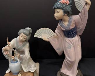 Lladró Porcelain Geisha Figurines – Flower Arranger and Fan Dancer A kneeling Lladró porcelain geisha arranging blossom branches in a vase on a low table, and a taller Lladró geisha in a pink kimono dancing with two open fans. Both have porcelain floral hair ornaments and detailed robes. An elegant pair for Lladro and Asian-themed figurine collectors.
