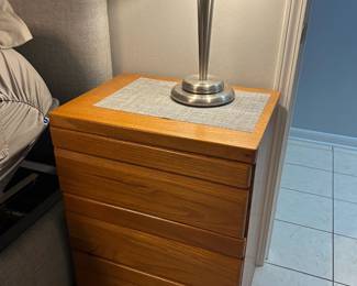 Wood Multi-Drawer Nightstand with Metal Dome Table Lamp A wood-grain nightstand with stacked drawers and a placemat on top, shown with a brushed metal table lamp with a frosted dome shade. A clean, simple bedroom piece. Confirm whether the lamp is included.