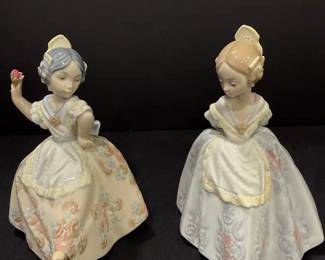 Lladró Porcelain Girls in Traditional Floral Gowns – Pair Two Lladró porcelain figurines of young women in full traditional gowns with lace-trimmed aprons, floral skirt patterns and high hair combs. One holds a single red flower. Delicate pastel painting makes this Lladro pair a graceful addition to a Spanish porcelain figurine collection.