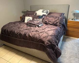 Sleep Number Bed with Plum Bedding and Two Nightstands A gray upholstered bed with a tall headboard, dressed in a plum pintuck comforter set with shams, pillows and extra bedding, flanked by two wood-grain nightstands with metal dome table lamps. A complete bedroom setup. Confirm the bed size in person.