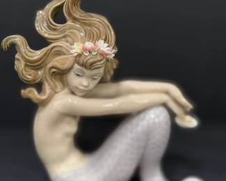 Lladró Porcelain Seated Mermaid with Flower Crown A Lladró porcelain mermaid figurine seated with her arms resting over her lavender-scaled tail, wearing a crown of sculpted porcelain flowers, with long flowing hair. A graceful Lladro mermaid figurine for any collection.
