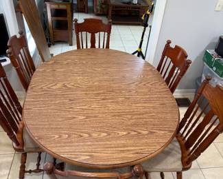 Oval Woodgrain Dining Table with Six Spindle-Back Chairs An oval dining table with a woodgrain top, shown with six spindle-back chairs with upholstered seats. A board that may be a table leaf is leaning against the wall behind it. A traditional dining set for a kitchen or dining room.