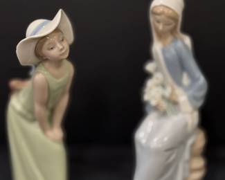 Lladró Porcelain Figurines – Girl in Sun Hat and Seated Lady with Flowers Two Lladró porcelain figurines in soft pastel colors: a young girl in a wide-brimmed sun hat bending forward, and a seated woman in a blue shawl and head covering holding white flowers. A collectible Lladro figurine pair for a display cabinet.