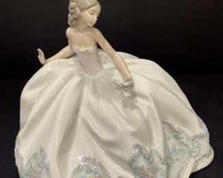 Glazed Figurine of a Lady in a Ball Gown A glazed ceramic figurine of a young woman seated in a full white ball gown, with one hand raised, an updo with a hair ornament, a rosebud at the bodice and pastel lace-textured floral trim at the hem. An elegant figurine centerpiece for a curio cabinet or collector's shelf.