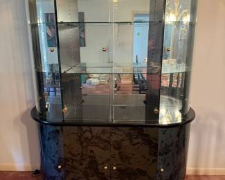 Lighted Curio Display Cabinet with Black Burl Base A large curio cabinet with curved glass sides, glass shelves, mirrored back panels and top lights, set on a black burl-pattern base with closed storage. A striking china or collectibles display cabinet. Lighting function not confirmed — sold as found.