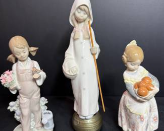 Three Lladró Porcelain Figurines – Girl Gardener, Hooded Girl with Staff and Girl with Oranges Three Lladró porcelain figurines: a girl in overalls holding flowers beside a watering can, a tall hooded girl with a staff on a round base, and a girl in a floral dress carrying a basket of oranges. Fine sculpted detail and soft colors. A strong lot for Lladro figurine collectors.