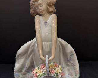 Glazed Figurine of a Girl with a Flower Basket A glazed ceramic figurine of a girl in a full pastel gown holding a basket of sculpted flowers, with windswept hair and a pink headband. The sculpted petals look crisp and colorful in the photo. A display-ready figurine for a shelf or collector's cabinet. Lladro