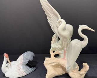 Lladró Porcelain Crane and Heron Bird Figurines A Lladró porcelain crane figurine resting with a red crown and spread wings, and a larger Lladró sculpture of two herons on a driftwood-style base, one with its wings raised high. Lladro bird figurines with fine feather detail for wildlife decor or a collector's shelf.