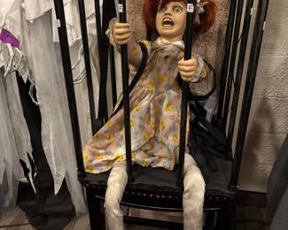 Caged Terror" Animated Crying Doll Prop red-haired doll trapped behind bars mounted to a high stool chair. A highly disturbing, premium character animatronic perfect for psychological horror displays. Features a distressed, vintage-style pigtail doll with piercing blue eyes, a classic duckling-print dress, and a terrifyingly realistic exposed teeth grimace. The character is completely enclosed within a custom faux-iron cage apparatus with integrated support straps, mounted atop a tall structural presentation stool to keep the scare right at eye level for your guests.