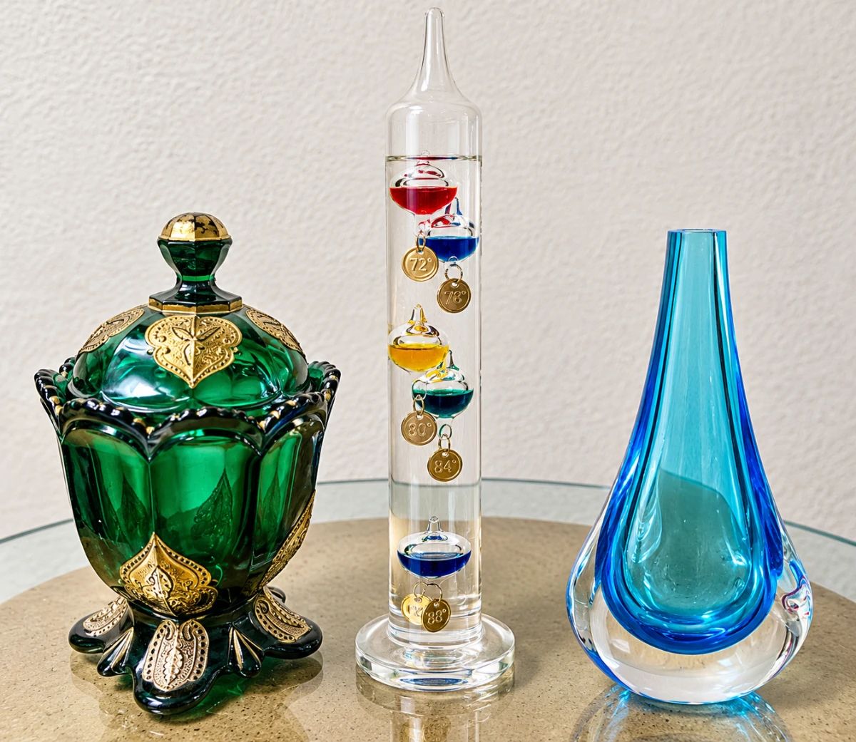 Emerald Green Decorative Glass Jar ◆ Rich green glass ◆ Ornate gold-tone embellishments ◆ Scalloped rim ◆ Matching decorative lid ◆ Footed base Galileo Glass Thermometer ◆ Tall clear glass cylinder ◆ Colorful floating glass bulbs ◆ Gold-tone temperature tags ◆ Decorative scientific instrument Blue Art Glass Vase ◆ Aqua and cobalt blue tones ◆ Teardrop shape ◆ Clear glass base ◆ Sculptural design ◆ Narrow opening