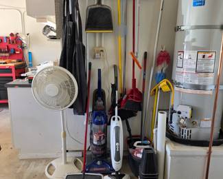 Collection of household cleaning supplies and garage items, including upright vacuums, a steam mop, a floor fan, brooms, a dustpan, and aprons