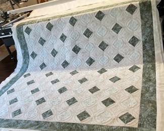 A construction-stage photograph of Cathy’s cream-and-sage quilt during longarm quilting, showing the repeating diamond layout, intricate circular stitching, and fabric extending beyond the unfinished edges.