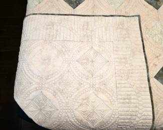 Cream-and-sage quilt folded back to reveal detailed quilting on the reverse. Circular motifs, diamond shapes, and closely spaced stitching give the pale fabric sculpted texture.
