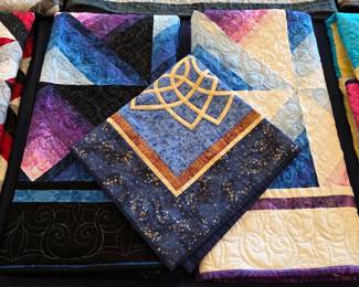 Blue, purple, cream, and black patchwork quilts displayed with a gold Celtic-knot motif piece. Close inspection reveals decorative quilting across the contrasting fabrics.