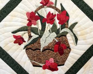 Floral basket appliqué featuring red blossoms, green leaves, and patterned basket fabrics. Radiating quilting frames the design within a dark green diamond border.