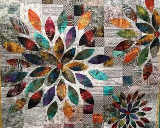 Floral appliqué art quilt with colorful petal shapes arranged over a patchwork background of text, music, and other printed fabrics. Decorative stitching adds detail throughout.