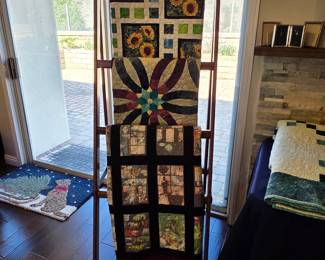 Handmade quilts displayed on a wooden ladder, featuring sunflower prints, a curved floral design, and contrasting framed patchwork blocks. A sampling of Cathy’s varied textile work.