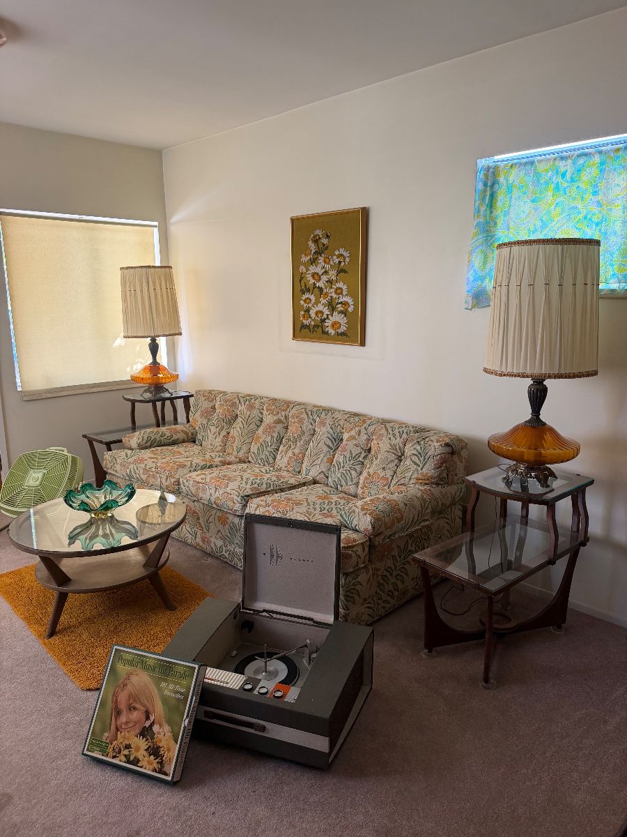 Charming 1960s living room featuring a vintage floral sofa, mid-century lamps, retro coffee table, record player, and nostalgic home décor.