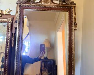 French Louis XVI–Style Carved Wood and Gilt Mirror with Elaborate Floral Crest — Rectangular mirror featuring an ornate openwork foliate crest, classical urn motif, carved garlands, decorative egg-and-dart molding, and hanging tassel ornaments. Rich gilt finish with contrasting reddish-brown accents. Traditional French Neoclassical design.