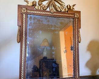 French Louis XVI–Style Carved Wood and Gilt Mirror with Ribbon Crest — Decorative rectangular mirror featuring an elaborate gilt ribbon-and-bow crest, floral corner rosettes, twisted-rope molding, and hanging foliate ornaments. Warm wood finish with gilded accents. Aged mirror plate with visible foxing and silvering loss.