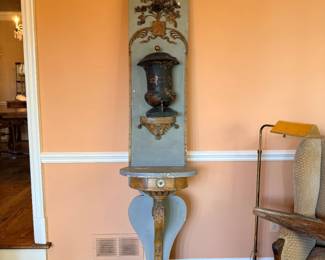 French Provincial–Style Painted Wall Fountain / Lavabo with Console Shelf — Unusual decorative wall fountain featuring a tall blue-gray painted wooden backboard with elaborate gilt floral and scrollwork embellishments. Centered with a dark painted metal lidded reservoir and spigot, above a carved ornamental shelf and narrow architectural support. Beautifully aged finish with decorative patina. A distinctive French Country accent piece.