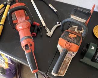 Selection of outdoor power tools and hand tools, including an orange electric hedge trimmer, a Stihl chainsaw, loppers, and a bow saw.
