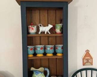 Green hutch topped with a kitty teapot and red floral teapots. Pitchers, cups and a cow creamer too. Moo!