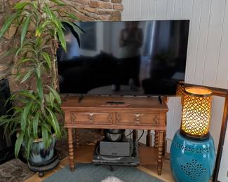 Vintage wood console table with spindle legs and two drawers, TV stand. Samsung flat screen TV with remote. Tall dracaena plant in blue glazed pot. Hanging golden pothos. Moroccan metal cylinder lamp. Turquoise ceramic garden stool/side table. Gray area rug.