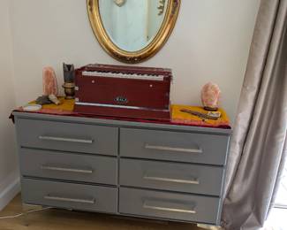 Gray 6-drawer dresser with silver bar pulls and chrome legs, modern. Gold oval beveled wall mirror, ornate vintage-style frame. Champagne/gold silk curtains (pair).