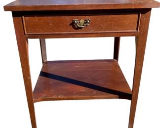 1017 - Mahogany Wood 1 Drawer Side Table (Description): Retro wood side table featuring a single drawer with a decorative brass pull handle. The table includes a lower shelf for additional storage and stands on tapered legs. Measures 29 inches tall by 23 inches wide by 19 inches deep.
