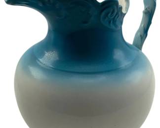 716 - Signed Pitcher 7"x7" (Description): A signed pitcher featuring a decorative handle and ruffled rim. The piece transitions in color from a darker blue at the top and base to a lighter shade in the middle. Measures 7 inches by 7 inches.