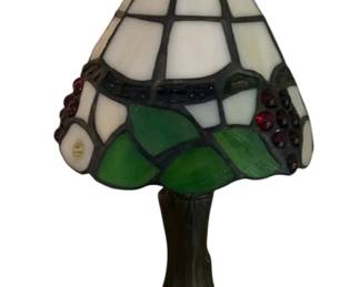 765 - Stained Glass Style Lamp 11" (Description): A stained glass lamp featuring a decorative shade with colorful glass panels in white, green, and deep red tones arranged in a grape and leaf motif. The shade is topped with a decorative finial and sits atop a bronzed metal base. Measures 11 inches in height. Used as-is electric/electronic/mechanical item. No guarantee all components present, untested if not otherwise stated.
