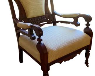 761 - Victorian Carved Upholstered Arm Chair (Description): Antique wood upholstered chair featuring carved wooden frame with decorative fretwork and scrolled arms. The chair is upholstered in a light-colored fabric and rests on turned legs with casters. Measures 34 inches high by 27 inches wide by 25 inches deep.
