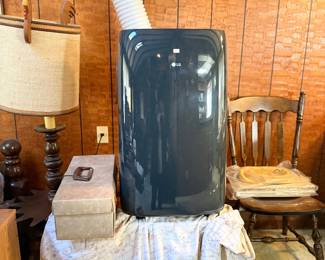 Modern dark grey LG portable air conditioner unit with exhaust hose, shown alongside a vintage table lamp and wooden side chair.