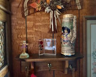 Rustic wall decor display featuring a wooden coat rack shelf holding a vintage ceramic German beer stein, acrylic Footprints prayer plaque, and a small crucifix, topped by a woven wicker fan swag with autumn florals.