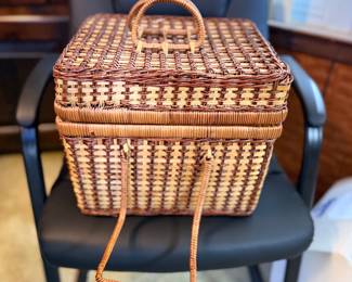 Vintage woven wicker sewing basket or picnic caddy featuring a top lid handle and a fold-down front handle, resting on a black chair.