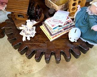 Rustic gear-shaped wooden side table holding a log cabin lamp, cookbooks, small pig figurines, and glassware.